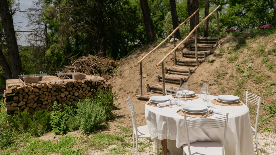 Tavolo rotondo all’aperto apparecchiato con tovaglia bianca, piatti e calici su un’area di ghiaia, circondato da verde. Sullo sfondo si vedono una scalinata in tronchi con corrimano in legno su un terrapieno, alberi e una catasta di legna con griglia.