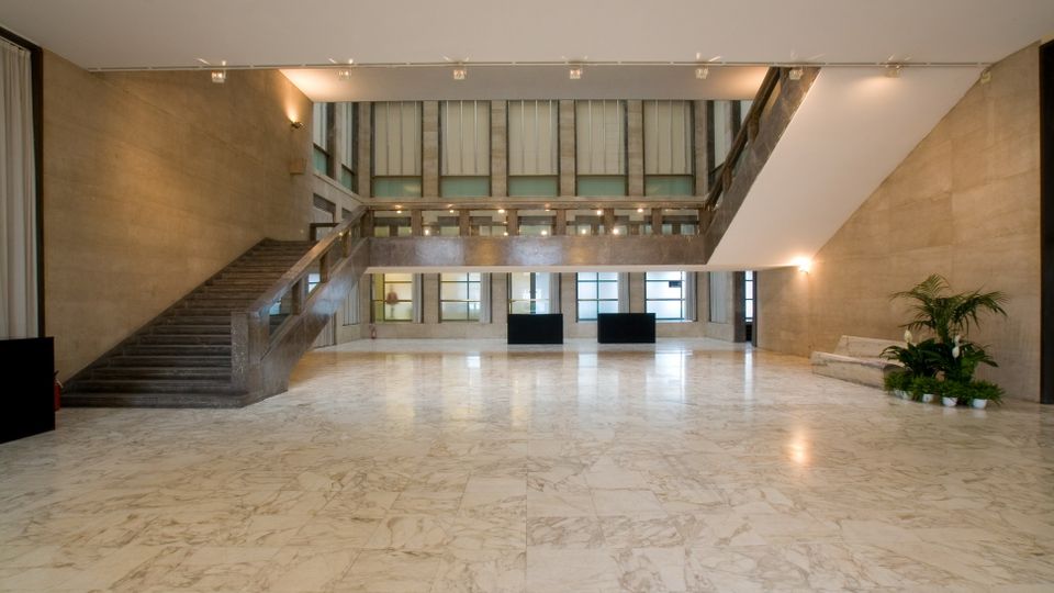 Ampio atrio interno di un edificio moderno con pavimento in marmo chiaro, pareti in pietra e grandi finestre sul fondo. A sinistra c’è una scala in pietra con corrimano, mentre a destra si vede un’area con piante e una panca, illuminata da luci a parete e a soffitto.