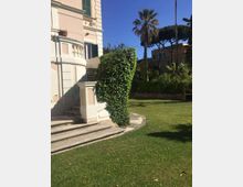Esterno di una villa in stile classico con facciata chiara, persiane verdi e scalinata curva con balaustra. In primo piano un prato curato con siepe potata e, sullo sfondo, palme e altri edifici tra la vegetazione sotto un cielo sereno.