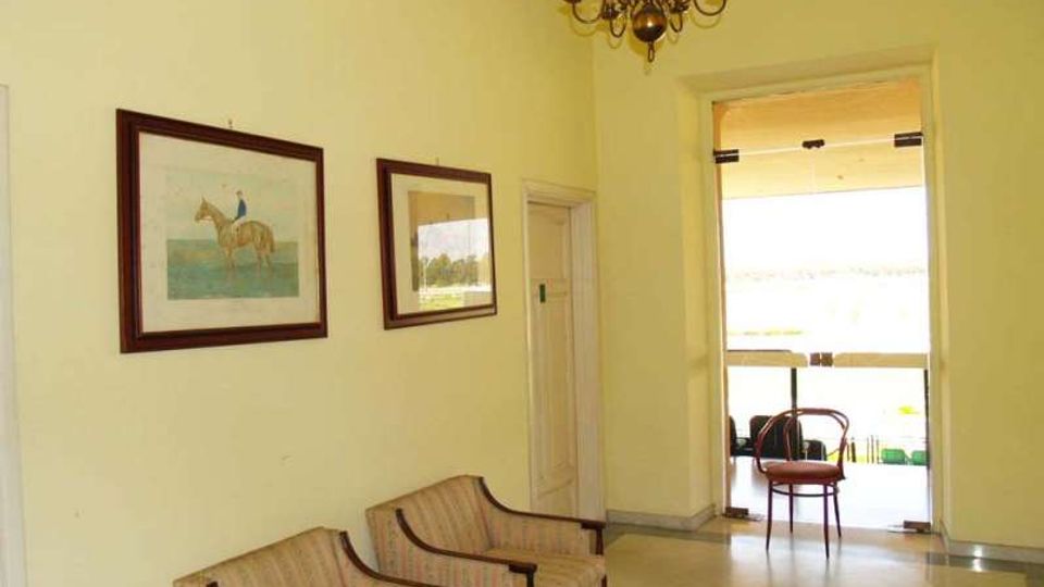 Interno di un corridoio o salottino con pareti chiare, due poltrone imbottite e quadri incorniciati appesi; dal soffitto pende un lampadario classico. In fondo, una porta-finestra aperta conduce a una veranda luminosa con sedie e vista sull’esterno.