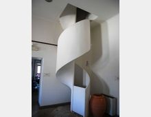 Scala a chiocciola in metallo verniciato bianco che sale verso un’apertura nel soffitto, collocata in un piccolo atrio interno con pareti chiare e pavimento in pietra. Accanto alla scala si vede un grande vaso in terracotta e una porta che conduce a un’altra stanza.