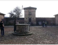 Cortile lastricato in pietra con un grande pozzo circolare in primo piano, dotato di bracci metallici per la carrucola. Sullo sfondo si vede un edificio in muratura con porta centrale e tetto a padiglione, affiancato da mura perimetrali e altri fabbricati.