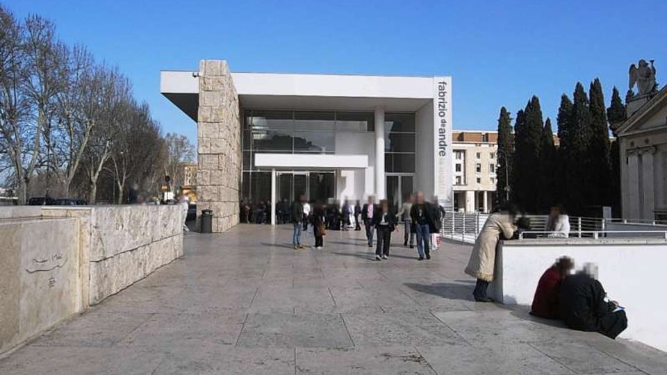 Ingresso di un edificio moderno con ampia facciata vetrata e tetto piano aggettante, affiancato da un pilastro rivestito in pietra e con la scritta verticale “fabrizio de andrè”. Davanti si apre una grande piazza pavimentata in pietra, con muretto laterale, alberi spogli e altri edifici sullo sfondo.