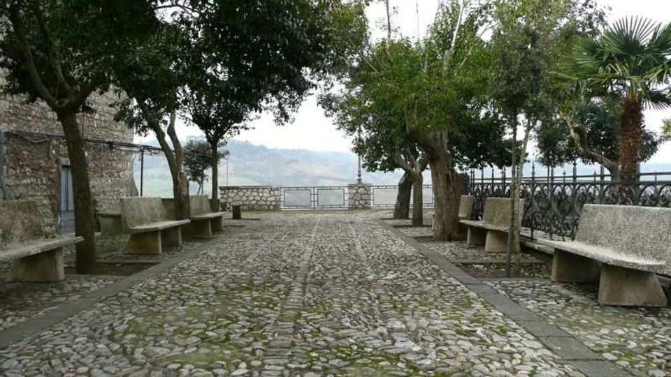 Passeggiata in ciottolato in un piccolo belvedere alberato, con panchine in pietra ai lati e ringhiera metallica sul bordo. Sullo sfondo si intravede un panorama collinare oltre un muretto in pietra.