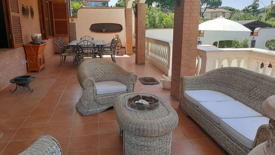 Ampio portico coperto di una villa con pavimento in cotto, colonne in mattoni e balaustra bianca, arredato con salotto in vimini e tavolo da pranzo in ferro. Sullo sfondo si vedono il giardino e alcune case, con un grande ombrellone nel cortile esterno.
