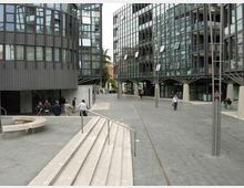 Piazza pedonale moderna pavimentata in pietra, con gradini e corrimano in primo piano e sedute in cemento. Ai lati si affacciano edifici contemporanei con grandi facciate vetrate e strutture metalliche, con lampioni alti lungo il percorso.