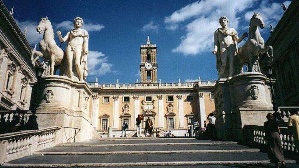 Scalinata monumentale che conduce a un palazzo storico con facciata simmetrica e torre dell’orologio centrale. Ai lati della scalinata ci sono grandi statue in pietra su piedistalli, con figure classiche e cavalli, sotto un cielo azzurro con nuvole.