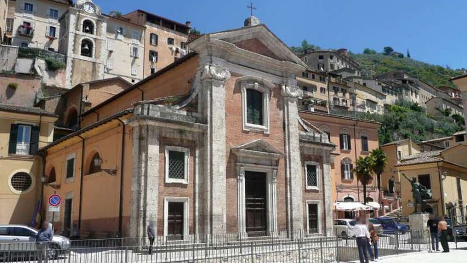 Facciata di una chiesa in mattoni e pietra con portale centrale e frontone triangolare sormontato da una croce, affacciata su una piazza con ringhiera. Sullo sfondo, case addossate a una collina e un campanile tra gli edifici.