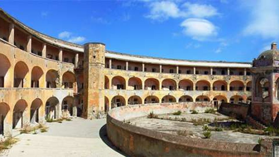 Cortile interno di un edificio storico a pianta semicircolare con due livelli di portici ad archi e pareti intonacate, affacciati su un’area centrale murata con vegetazione. Sul lato destro si vede una piccola struttura ottagonale con cupola e decorazioni, sotto un cielo azzurro con nuvole.