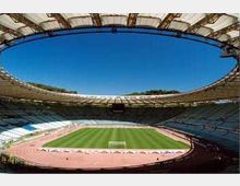 Veduta interna di un grande stadio con copertura ellittica e gradinate che circondano un campo da calcio in erba, con pista di atletica rossa tutt’intorno. Il cielo azzurro è visibile attraverso l’ampia apertura centrale del tetto.