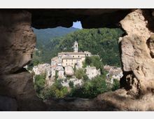 Veduta di un borgo collinare con case in pietra e un campanile centrale, incastonato tra boschi e montagne. Il paesaggio è inquadrato da un’apertura irregolare in un muro di pietra in primo piano.