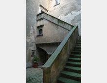 Cortile interno di un edificio storico con scala esterna in pietra che sale lungo un alto muro intonacato e in parte scrostato, con piccolo balcone e finestre. Alla base si vedono un arco di passaggio, pavimentazione in mattoni a spina di pesce e un vaso con pianta.