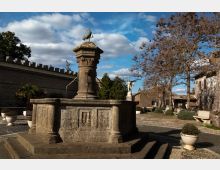 Fontana monumentale in pietra al centro di una piccola piazza lastricata, con colonna decorata e scultura in cima. Intorno si vedono un muro merlato, alberi spogli, panchine e edifici storici sotto un cielo azzurro con nuvole.