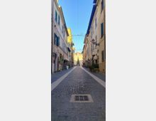 Strada stretta in un centro storico con pavimentazione in sampietrini, fiancheggiata da edifici a più piani con persiane e lampioni a parete. In alto sono appese luminarie a forma di stella e, in fondo alla via, si vede una piazza con un grande edificio e un albero decorato.