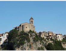 Chiesa in pietra con campanile su una sommità rocciosa, affiancata da un borgo di case addossate alla scogliera. Vegetazione arbustiva sul pendio e cielo sereno sullo sfondo.
