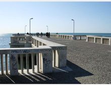 Molo sul mare con ampia pavimentazione in pietra e parapetti in cemento segnati da graffiti, che si estende verso l’orizzonte. Lungo il percorso sono presenti alti lampioni e si vede il mare aperto su entrambi i lati.