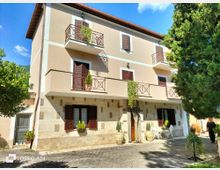 Facciata di una palazzina residenziale a tre piani con intonaco beige e base in pietra, persiane in legno scuro e balconi con ringhiere metalliche e piante ricadenti. Davanti c’è un cortile pavimentato con vasi e un piccolo set da esterno, circondato da alberi sotto un cielo azzurro.