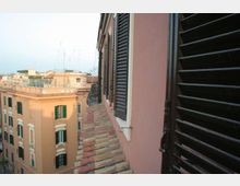 Vista da una finestra su un tetto inclinato in tegole e sulla facciata di un edificio color rosa con persiane nere. Sullo sfondo si vedono palazzi urbani color ocra con finestre e terrazze, sotto un cielo chiaro.