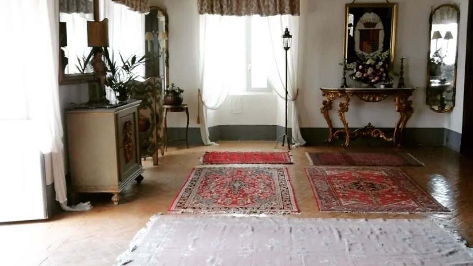 Salone interno in stile classico con soffitto decorato a cassettoni, grandi finestre con tende leggere e pavimento in parquet a spina di pesce. La stanza è arredata con consolle intagliate, specchi e tappeti persiani disposti sul pavimento.