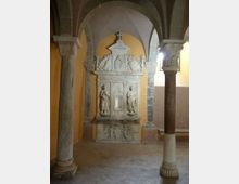 Interno di una chiesa o cappella con volte a crociera e colonne in pietra, pavimento in pietra e pareti color ocra. Al centro si vede un altare o monumento marmoreo riccamente scolpito con rilievi e figure, incorniciato da archi e pilastri.