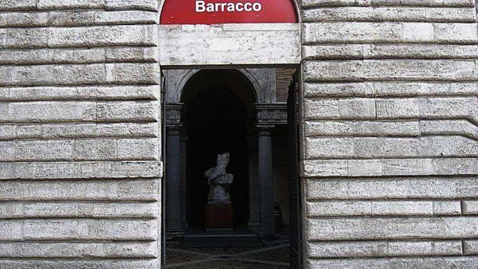 Ingresso del Museo Barracco con portale ad arco in pietra bugnata e insegna rossa sopra l’architrave. All’interno del corridoio si intravede una statua, con pavimentazione in lastre di pietra e mattoni davanti all’entrata.