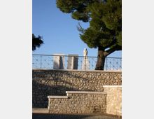 Terrazza rialzata con muri in pietra e ringhiera metallica, con una statua su piedistallo e grandi blocchi rettangolari sullo sfondo. Un grande albero sempreverde domina il lato destro contro un cielo sereno.
