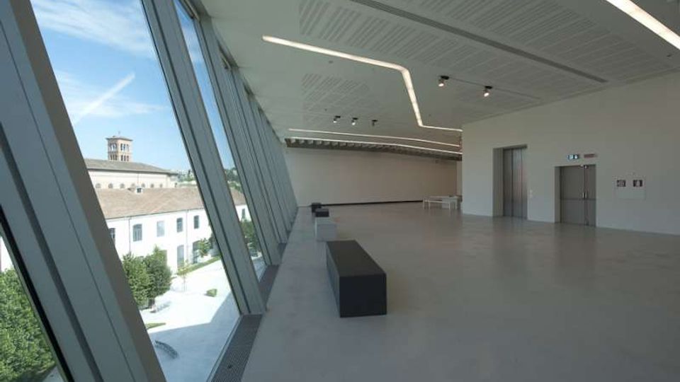 Ampio interno di edificio moderno con pavimento chiaro, soffitto con illuminazione lineare e grandi vetrate inclinate a tutta altezza sul lato sinistro. Dalle finestre si vede un complesso storico con cortile e campanile; lungo la sala sono presenti una panca scura e pareti bianche con porte.