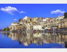 Borgo collinare con case in pietra e facciate chiare addossate al pendio, affacciato su un lungolago con piccole strutture portuali. In primo piano l’acqua calma riflette gli edifici e il cielo azzurro con alcune nuvole.