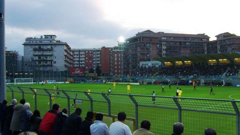 Stadio di calcio con campo in erba e recinzione metallica in primo piano, visto dalle gradinate con una tribuna coperta illuminata sul lato opposto. Sullo sfondo si vedono palazzi residenziali e un cielo nuvoloso al crepuscolo.
