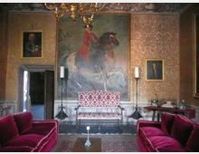 Salone interno in stile storico con pareti rivestite di carta da parati decorata, grande dipinto equestre centrale e ritratti incorniciati ai lati. Al centro una panca imbottita e due alti candelabri, con divani in velluto rosso in primo piano e lampadario sospeso al soffitto.