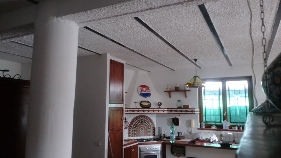 Cucina abitabile con soffitto a travi scure a vista, pensili in legno e piano cucina angolare con forno e piastrelle decorative. Una finestra con vetri verde-azzurri illumina la zona lavabo, con lampada a sospensione sopra il bancone.