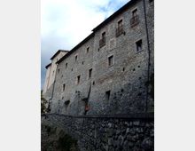 Grande edificio storico in pietra su un pendio, con facciata lunga e irregolare, piccole finestre e balconcini in ferro battuto. In primo piano si vede un muro di contenimento in pietra lungo una strada con lampioni, sotto un cielo nuvoloso.