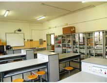 Aula laboratorio scolastico con banchi da lavoro e sgabelli gialli, illuminazione a neon e una lavagna bianca con proiettore a parete. Sul lato destro ci sono armadi vetrati con attrezzature e strumenti, e un banco con lavello sul fondo.
