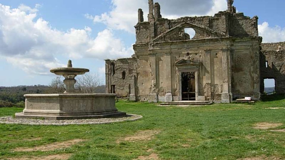 Rovine di un edificio storico in muratura con facciata parzialmente crollata e grande apertura centrale, situato in un prato. In primo piano si vede una fontana in pietra circolare, con cielo parzialmente nuvoloso sullo sfondo.
