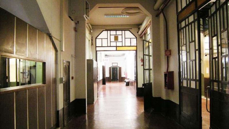 Corridoio interno di un edificio, lungo e stretto, con pavimento in legno e soffitto alto. Ai lati ci sono pareti con finestre e porte vetrate a riquadri metallici, e in fondo un grande infisso vetrato che lascia entrare luce.