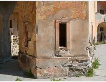 Facciata di un edificio antico e degradato, con intonaco scrostato e muratura in pietra a vista, e una finestra rettangolare senza vetro. L’edificio si affaccia su una strada lastricata, con archi e altri muri in rovina sullo sfondo.