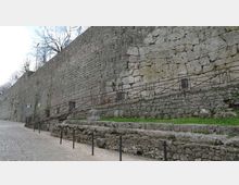 Lungo muro di cinta in pietra con tessitura irregolare, affiancato da una strada lastricata e da terrazze in muratura a gradoni con piccoli tratti d’erba. Una rampa laterale con parapetto metallico costeggia il muro, con alberi spogli visibili sopra la sommità.