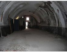 Interno di un grande tunnel o rifugio sotterraneo con volta a botte in cemento, pareti scrostate e pavimento polveroso con detriti. In fondo si vede un’apertura rettangolare verso l’esterno e alcune grandi porte o pannelli appoggiati lungo il lato sinistro.
