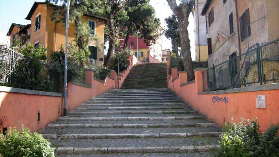 Ampia scalinata in pietra che sale tra muretti intonacati color salmone, con ringhiere e lampioni lungo i lati. Ai lati della scalinata si vedono case residenziali dai toni caldi e alberi alti che ombreggiano il percorso.