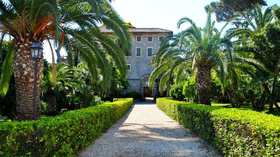 Viale in ghiaia fiancheggiato da siepi potate e alte palme in un giardino mediterraneo, che conduce a una villa storica in pietra con tetto in tegole e arco d’ingresso centrale. Vegetazione rigogliosa e lampioni classici lungo il percorso.