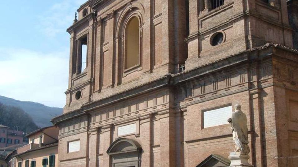 Facciata di una grande chiesa in mattoni con portali scuri, finestre ad arco e campanile laterale a loggia, vista da una piazza. In primo piano si vede una statua su piedistallo, con edifici residenziali e colline sullo sfondo.