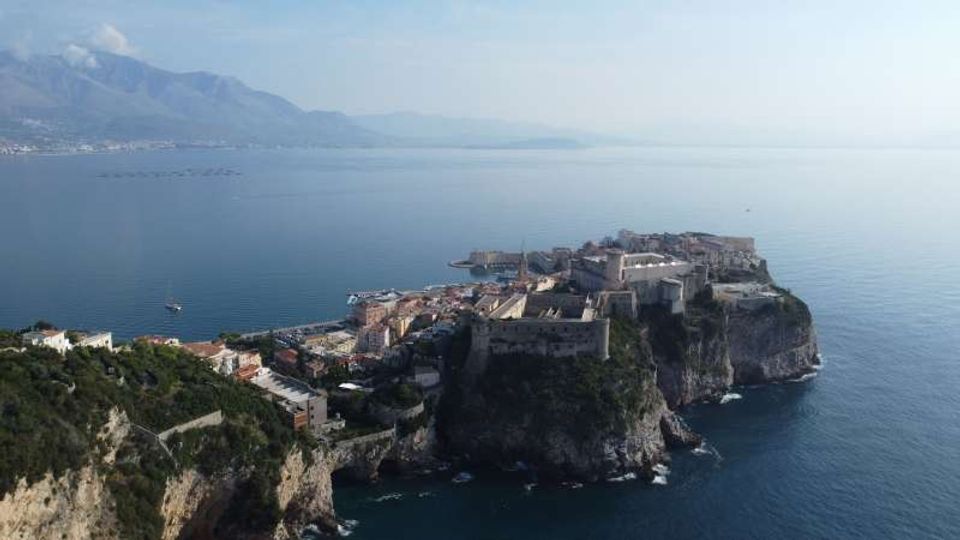 Veduta panoramica di una cittadina costiera con edifici storici e fortificazioni su un promontorio roccioso a picco sul mare. In lontananza si vedono montagne e una costa che si estende lungo l’orizzonte, con acque calme intorno al promontorio.