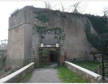 Ingresso ad arco in una massiccia fortificazione in muratura di mattoni e pietra, parzialmente coperta da vegetazione, raggiunto da un vialetto su un piccolo ponte con muretti laterali. Intorno si vede un’area verde con alberi spogli e pendii erbosi.