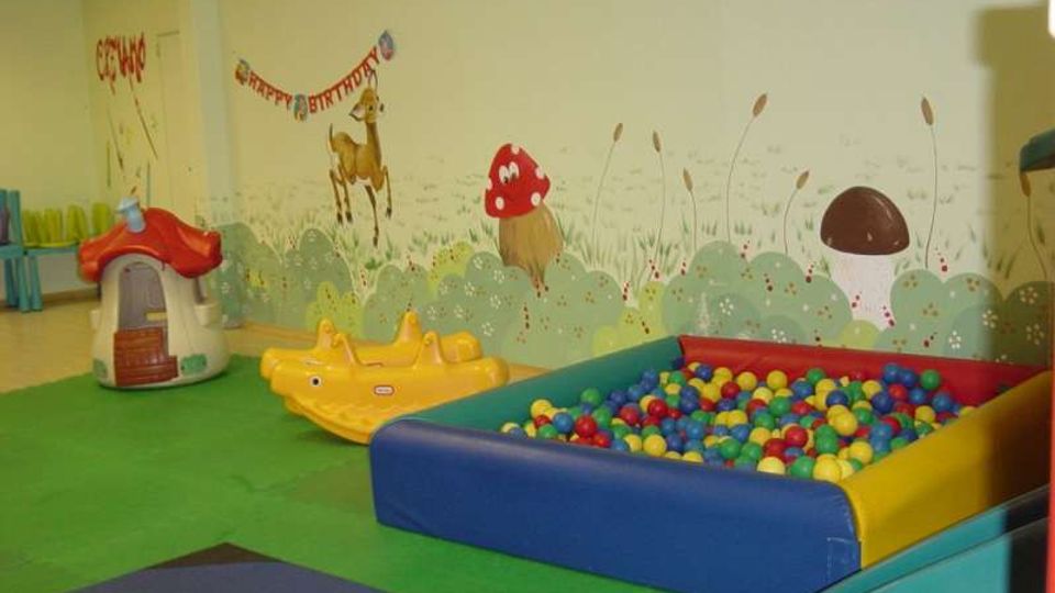 Area giochi interna per bambini con pavimento morbido verde, piscina di palline colorate imbottita e piccoli giochi in plastica. Parete decorata con murales a tema natura e compleanno, con funghi, animali e scritta “Happy Birthday”.