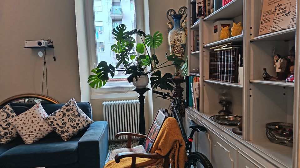 Salotto domestico con divano blu e cuscini decorati, poltrona con coperta e pavimento a piastrelle con motivi geometrici. Vicino a una finestra con radiatore e grande pianta in vaso, si vedono una libreria alta piena di libri e oggetti e una bicicletta appoggiata.