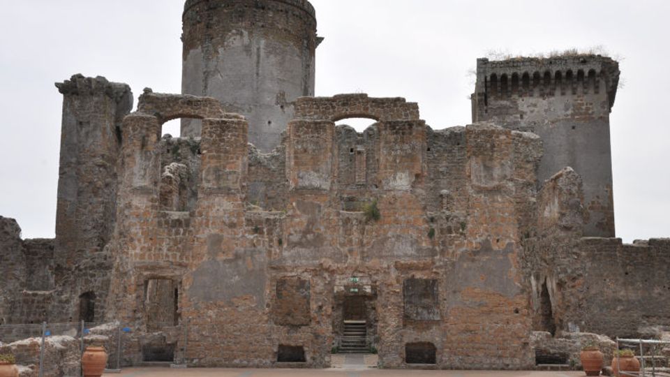 Rovine di un castello in pietra e mattoni con muri parzialmente crollati, archi e aperture murate, dominato da una grande torre cilindrica e da una torre quadrata sullo sfondo. In primo piano si vede un cortile pavimentato con vasi e una scalinata centrale che conduce all’interno.
