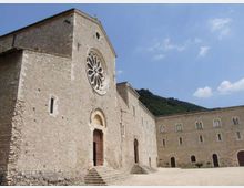 Facciata di una chiesa in pietra chiara con grande rosone circolare e portale ad arco raggiunto da alcuni gradini, affacciata su un ampio cortile. Sul lato destro si estende un edificio in pietra con finestre regolari e archi al piano terra, con colline verdi e cielo azzurro sullo sfondo.