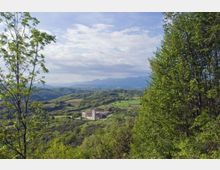 Vista panoramica su una valle collinare verde con campi e boschi, con un complesso edilizio in pietra su un crinale al centro. Alberi in primo piano incorniciano il paesaggio, con montagne lontane sotto un cielo parzialmente nuvoloso.