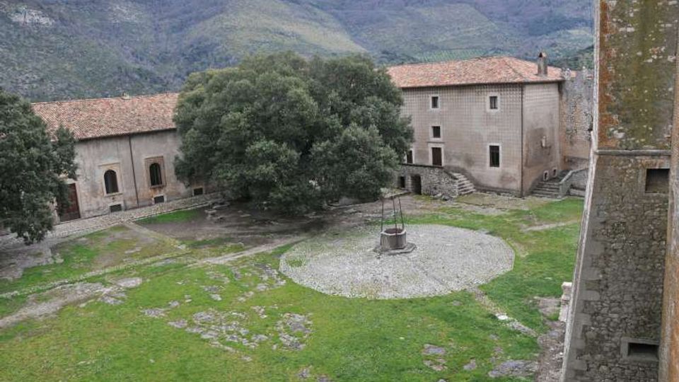Cortile erboso di un complesso in pietra con edifici dai tetti in tegole, un grande albero centrale e un pozzo circolare su un’area di ghiaia. Sullo sfondo si vedono colline e montagne boscose.