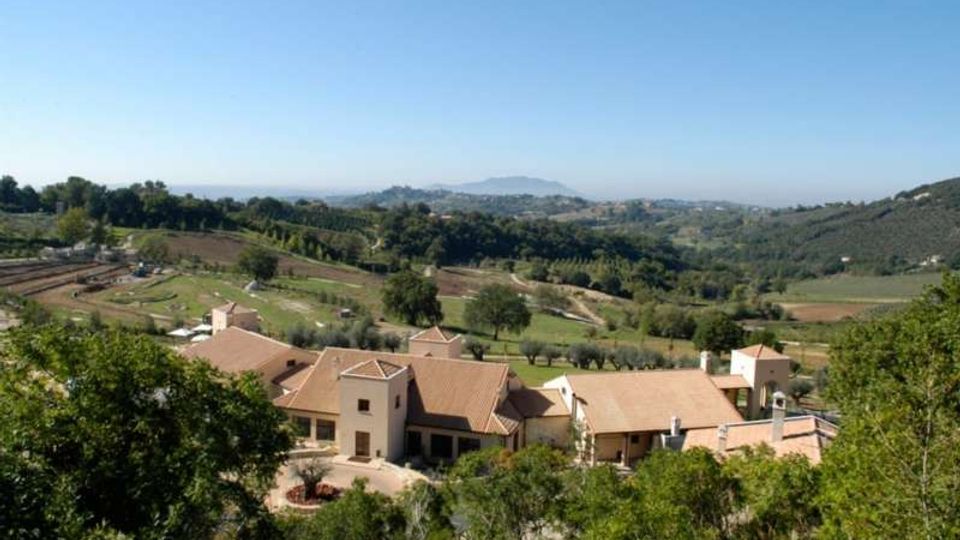Veduta panoramica di un complesso di edifici rurali con tetti in tegole color terracotta, circondato da alberi e strade sterrate. Sullo sfondo si estendono colline coltivate e campi verdi fino a montagne lontane sotto un cielo sereno.